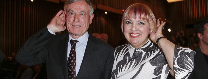 Horst Köhler und Claudia Roth beim 14. Deutschen Hörfilmpreis am 15. März 2016. Sie stehen im dunklen Kinosaal und halten jeweils eine Hand hinter ihr Ohr.