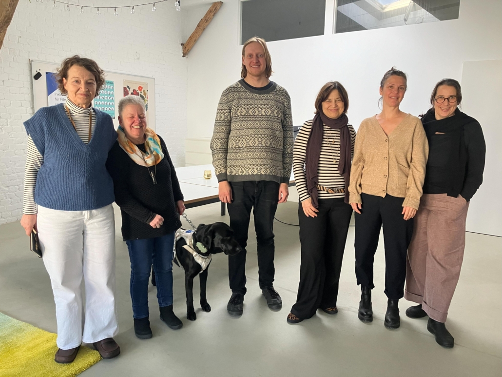 Auf dem Foto stehen sechs Menschen und ein Hund bei einem Vereinstreffen in einem hellen, modern eingerichteten Raum. 
Es sind: Susanne Willnow, Andrea Eberl mit ihrem schwarzen Labrador Frodo im Führgeschirr, Felix Koch, Christine Heimannsberg und Jutta Polić.
Sie lächeln in die Kamera. 
Susanne Willnow (ehemaliges Vorstandsmitglied) hat dunkelblondes, lockiges Haar, das ihr bis unter die Ohren reicht. Susanne trägt einen blauen, ärmellosen Strick-Pullunder mit V-Ausschnitt, darunter ein weißes Rollkragen-Longsleeve mit schwarzen Querstreifen. Zu ihrer weißen, weiten Hose trägt sie dunkelbraune Schuhe.
Andrea Eberl (ehemaliges und neu gewähltes Vorstandsmitglied) hat graues Haar im Igelschnitt. Andrea trägt einen schwarzen Strickpullover mit einem gemusterten Schal in Beige-Gelb-Türkis-Tönen, darüber hängt eine Hornpfeife. Sie hat dunkelblaue Jeans und schwarze Schuhe an und hält die Leine ihres Hundes.
Felix Koch (neu gewähltes Vorstandsmitglied) ist rotblond. Sein glattes Haar ist Nackenlang. Felix trägt einen weiten hellgrauen Pullover mit Norweger-Muster, schwarze Jeans und schwarze Schuhe.
Elisabeth Löhmann (ehemaliges Vorstandsmitglied) ist brünett. Ihr stufig geschnittenes Haar ist kinnlang. Elisabeth hat ein schwarz-weiß gestreiftes Longsleeve mit einer schwarzen Hose und Schuhen in Leopardenmuster kombiniert. Dazu trägt sie einen braunen Schal und einen schmalen gestreiften Gürtel.
Christine Heimannsberg (ehemaliges Vorstandsmitglied) trägt ihr Haar hochgebunden. Christine hat ein beiges, geknöpftes Cardigan und eine schwarze, kürzer geschnittene Hose und schwarze Boots an.
Jutta Polić (neu gewähltes Vorstandsmitglied) trägt eine runde Hornbrille und ihr braunes Haar zum Pferdeschwanz gebunden. Jutta hat einen schwarzen Pullover und eine rosé-farbene, weite Hose an. Dazu trägt sie schwarze Schuhe.
Der Raum hat weiße Wände, sichtbare Holzträger und eine große Fensteröffnung an der Decke. Im Hintergrund sieht man ein Whiteboard mit farbigen Plakaten sowie einen großen Tisch.
Über der Gruppe hängen Lichterketten.