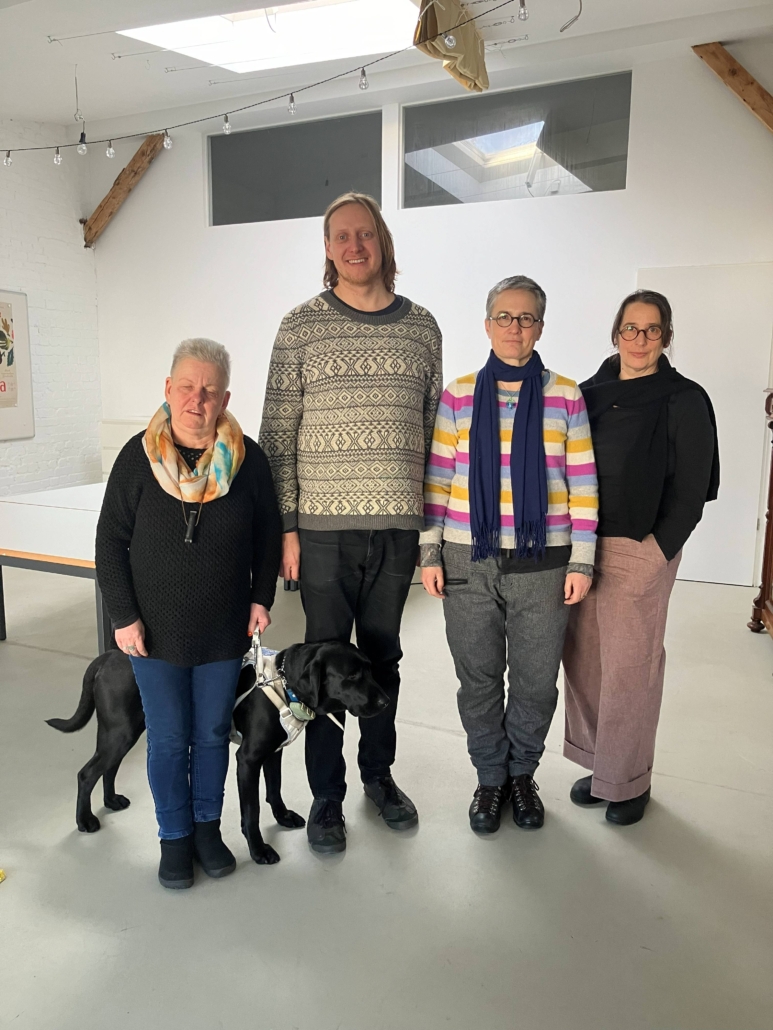 Auf dem Foto stehen vier Erwachsene und ein Hund in einem hellen, modern eingerichteten Raum. Alle blicken in die Kamera und wirken freundlich und entspannt.
Es sind: Andrea Eberl mit ihrem schwarzen Labrador Frodo im Führgeschirr, Felix Koch, Annabelle Wick und Jutta Polić.
Andrea Eberl (ehemaliges und neu gewähltes Vorstandsmitglied) hat graues Haar im Igelschnitt. Andrea trägt einen schwarzen Strickpullover mit einem gemusterten Schal in Beige-Gelb-Türkis-Tönen, darüber hängt eine Hornpfeife. Sie hat dunkelblaue Jeans und schwarze Schuhe an und hält die Leine ihres Hundes.
Felix Koch (neu gewähltes Vorstandsmitglied) ist rotblond. Sein glattes Haar ist Nackenlang. Felix trägt einen weiten hellgrauen Pullover mit Norweger-Muster, schwarze Jeans und schwarze Schuhe.
Annabelle Wick hat kurze graumelierte Haare und trägt eine runde Brille. Annabelle trägt einen mehrfarbigen Wollpullover mit Streifen in Rosa, Gelb, Blau, Grau und Weiß. Sie hat einen blauen Schal um. Zu ihrer grauen Hose trägt sie schwarze Wanderstiefel.
Jutta Polić (neu gewähltes Vorstandsmitglied) trägt eine runde Hornbrille und ihr braunes Haar zum Pferdeschwanz gebunden. Jutta hat einen schwarzen Pullover und eine rosé-farbene, weite Hose an. Dazu trägt sie schwarze Schuhe.
Der Raum hat weiße Wände, sichtbare Holzträger und eine große Fensteröffnung an der Decke. Im Hintergrund sieht man ein Whiteboard mit farbigen Plakaten sowie einen großen Tisch. Über der Gruppe hängen Lichterketten.